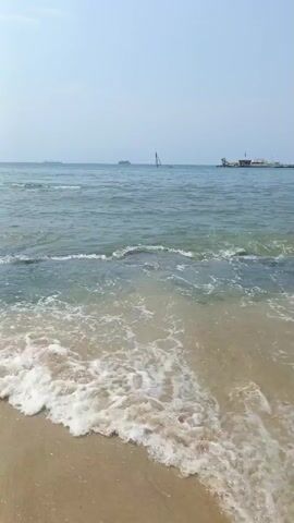 The video shows a beach scene with gentle waves washing onto the sandy shore. In the distance, several ships are visible on the horizon under a light sky. As the video progresses, a group of people are seen swimming in the water. The scene shifts from a bright, clear sky to a more overcast and cloudy atmosphere.