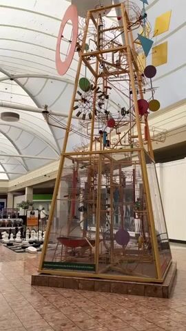 A tall, complex marble run sculpture with colorful geometric shapes and spinning wheels stands in the center of a spacious, well-lit indoor shopping mall. The sculpture is enclosed in clear glass panels at its base. In the background, shoppers walk along a wide tiled corridor lined with storefronts, including a JCPenney. The mall ceiling is a translucent white fabric structure supported by a metal frame. The lighting is bright and even, suggesting daytime.