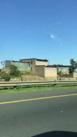 The video shows a view from a moving vehicle driving on a road. Beside the road, on the right, are several structures that appear to be dwellings constructed from basic materials. A green guardrail runs along the roadside. The sky is a clear, bright blue. A road sign indicating lane merging is visible, followed by a billboard with advertising on it. Vegetation is visible growing around the structures.