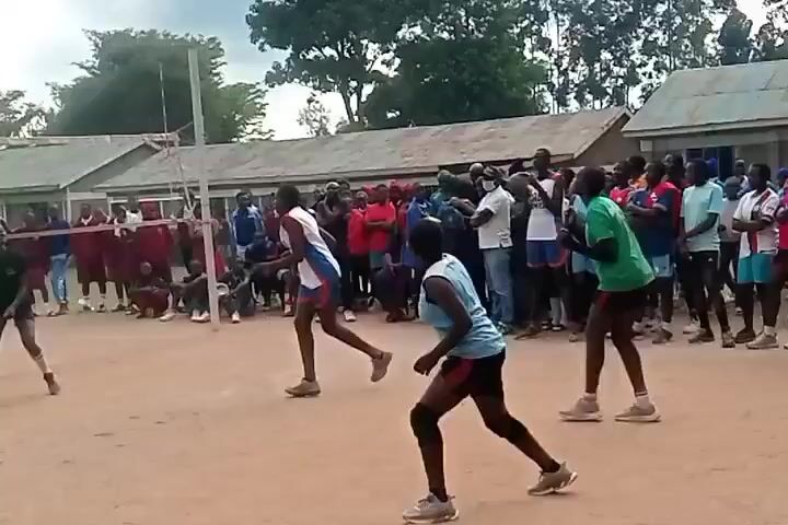 A volleyball game is in progress. Several players are on each side of the net, and a crowd of spectators is watching. The players are wearing different colored uniforms. The game is being played on a dirt court. The weather is overcast. There is a building in the background. The ball is in the air, and players are jumping and reaching for it.
