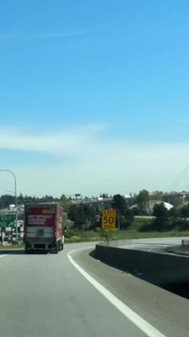The video depicts a view from a car driving on a highway on a clear, sunny day. In front of the car, a McDonald's delivery truck is driving along the same road. The truck's rear has an advertisement stating "Not Without Canadian Farmers".  An exit sign indicating "Exit 50 km/h" is visible on the right.  In the background, a large cable-stayed bridge is visible. The environment consists of typical highway infrastructure, including the road, safety barriers, and patches of greenery.