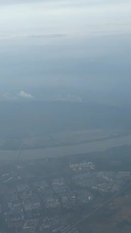 The video shows an aerial view of a cityscape under an overcast sky. In the distance, a river winds through the landscape, with mountains visible behind it. A bridge spans the river, and clusters of buildings form a dense urban area. The air appears hazy, and the colors are muted due to the cloudy weather.