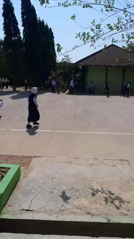 The video shows a group of students, possibly in a schoolyard. Several students are wearing dark uniforms with white headscarves, suggesting they might be female students. One student in a dark uniform and white headscarf is running towards the left side of the frame, followed by another student in a similar outfit. In the background, other students are visible, some running or standing near a green building with a brown roof. The ground is paved, and there are trees and a blue sky visible. The overall impression is of children playing or participating in an activity outdoors.