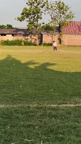 A boy in a white t-shirt and black shorts kicks a red ball on a grassy field. He is in the middle of the field, and the ball is in the air. In the background, there are trees and buildings. The sun is shining, and there are shadows on the grass. The boy is the only person visible.