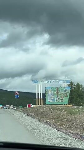 A short video shows a view from a car as it drives on a road. The car passes by a village nestled in a valley with a dense forest in the background. The sky is cloudy with dark, dramatic clouds. On the right side of the road, there is a large sign with Cyrillic lettering and a painting depicting people in traditional clothing. There are also road signs, including a warning sign for a steep incline and a circular sign with a blue background and a white arrow. The village has houses with colorful roofs. Another car is seen driving away from the camera on the road.
