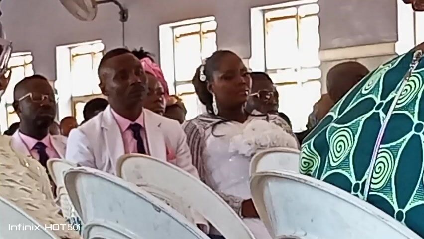 The video shows a scene in what appears to be a church, with a seated congregation. The focus is on a couple sitting in the front row, possibly the bride and groom, as they are dressed in white. The man wears a white suit jacket, and the woman wears a white dress with what looks like white flowers.  Behind them and around them, other people are seated on white plastic chairs. Some attendees are wearing glasses.  Several women are wearing elaborate headwraps. The room has multiple windows. Music is playing in the background. The camera is positioned at an angle, showing the side of the congregation. The footage appears to be recorded on a mobile device, as indicated by the watermark "Infinix HOT 50i" in the lower left corner of the frame.