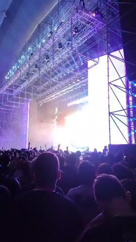 The video shows a concert with a large crowd. The stage is brightly lit with a massive lighting rig overhead. Some concert-goers are holding up their phones, presumably filming or taking pictures. The ceiling of the venue is visible and has a distinct curved pattern with several lights. The color of the lights shifts between blue and pink.