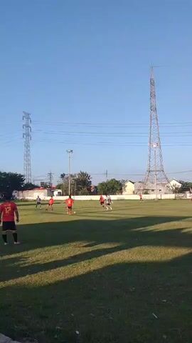 The video displays an outdoor scene of a soccer game played on a green grass field under a clear blue sky. Approximately ten to twelve individuals are visible, running and moving across the field, engaged in playing soccer. One player in the foreground, seen from behind, wears a red and black jersey with the number "12." Other players are further away, some wearing red or orange shirts and others in white or light-colored shirts. Long shadows are cast across the grass, suggesting either morning or late afternoon. In the background, two distinct metal towers dominate the skyline: a tall, slender lattice tower on the right, and a larger, broader power transmission tower with multiple overhead power lines on the left. Trees and some residential or industrial buildings are visible in the distant background. A single light pole stands in the mid-ground.