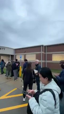 The video shows a line of people standing outside a building labeled "DIV. OF PUBLIC SAFETY DRIVER LICENSE". The sky is overcast. Most of the people are facing away from the camera, towards the entrance of the building. Some are looking at their phones. There are yellow parking barriers lining the front of the building and handicapped parking spots nearby. The building is made of a combination of brick and light-colored stone. Some windows are visible.