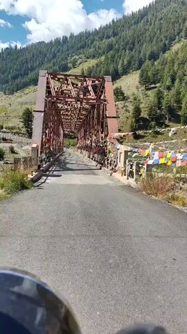 The video shows a POV shot from a vehicle, likely a motorcycle, approaching and crossing a large, rusty, metal truss bridge. The bridge is adorned with numerous colorful prayer flags strung along its sides. The road leading to and across the bridge is paved and appears to be in good condition. The surrounding environment is mountainous, with steep slopes covered in lush green trees and patches of bare earth. The sky is partly cloudy, indicating fair weather. The sun is casting distinct shadows across the bridge, highlighting the structure's intricate metalwork. The perspective suggests a steady forward movement across the bridge, with no other vehicles or pedestrians immediately visible on the bridge itself.