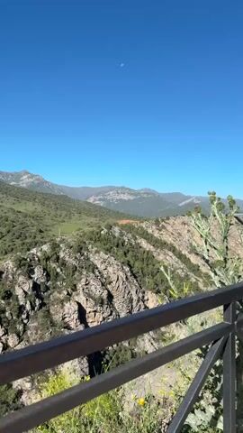 The video captures a panoramic view of a mountainous landscape under a clear blue sky. The foreground features a dark metal railing, partially obscured by green foliage and yellow wildflowers. Beyond the railing, a steep, rocky gorge descends, with trees and vegetation clinging to its slopes. The midground showcases rolling green hills dotted with shrubs and scattered trees. In the distance, a range of larger, more rugged mountains stretches across the horizon, their peaks partially covered in trees. A faint crescent moon is visible in the upper right portion of the sky. The overall impression is one of a vast, natural environment bathed in sunlight.