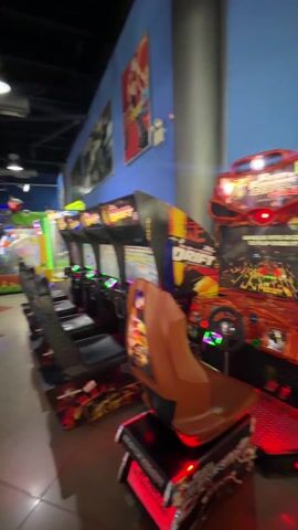 The video shows the interior of an arcade. A line of racing video game machines is visible, one with a "DRIFT" logo. A man in a red shirt is playing a dance-style arcade game. Other people, including a child, are standing nearby. The arcade has various game machines. The floor is tiled in a diamond pattern. The ceiling is black with visible ductwork and lighting fixtures.