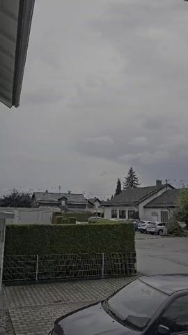 The video shows an exterior view of a suburban neighborhood under a cloudy sky. In the foreground, the hood and windshield of a dark-colored car are visible. Behind the car is a paved area with what appears to be a brick pattern. A low hedge runs across the middle ground, bordered by a dark fence. Beyond the hedge, several houses with dark roofs are visible, along with parked cars. Tall evergreen trees are scattered throughout the scene, and the sky is filled with thick, gray clouds.