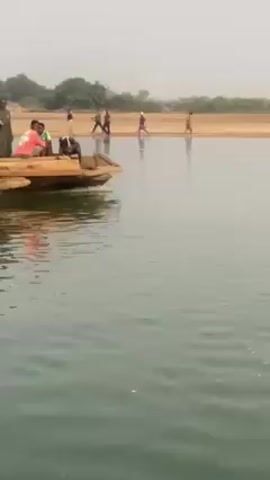 The video shows a makeshift raft carrying people and a red car across a body of water. The raft is made of logs. The water appears calm. People are standing and sitting on the raft alongside the car. The sky is overcast. There is land visible on the other side of the water. The overall impression is that this is a rudimentary form of transportation in a rural area.