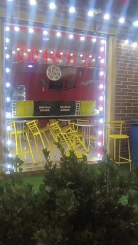 A video of a storefront at night. The storefront has a large window with a red interior wall, a HALAL logo, and a phone number visible. The window is framed by white lights. Inside the store, there are several yellow tables and chairs, some of which are stacked or askew. There are also yellow signs with black text that read "DOVRAN DONER". In front of the storefront is a dark green bush. To the right of the storefront, a blue barrel is visible.