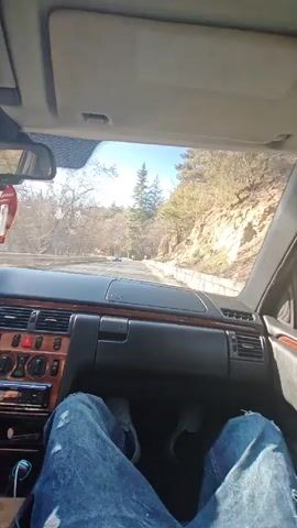 A driver is navigating a car along a winding road surrounded by trees. The video is shot from the passenger seat, showing the driver's legs in blue jeans, the car's dashboard with wood trim, and the road ahead. The environment features trees and rocky hillsides. The weather appears sunny, with a bright sky visible. The camera remains stationary throughout the video.