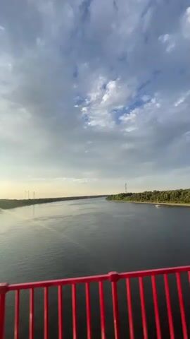 The video shows a wide river with a boat on the right side. The water is dark and reflects the sky, which is filled with clouds and sunlight. There is a red railing in the foreground, suggesting the video is taken from a bridge. The riverbanks are lined with trees, and there are some structures visible in the distance on the left bank. The overall scene is a landscape view of a river under a cloudy sky.