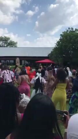 A crowd of people dances on a grassy lawn, moving rhythmically as captured in a handheld video. Individuals dressed in colorful attire, including pink, yellow, orange, and floral patterns, face away from the camera and toward a building, possibly a reception venue, with a gray roof. A red umbrella stands prominently near the building. The camera holder, positioned in the front of the crowd, also participates in the event while recording. The sky above features fluffy white clouds against a blue backdrop. A tree is visible on the right side of the frame.