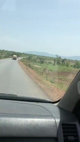 The video captures a first-person perspective from inside a moving vehicle, looking forward through the windshield on a clear, sunny day. The scene depicts a drive along an asphalt road in what appears to be a rural or semi-rural setting, consistent with the location context of Bokana, Nigeria.

In the foreground, parts of the vehicle's dark dashboard, the bottom edge of the windshield, and a light-colored side pillar are visible. The road ahead is paved, with visible white lane markings. A large, covered truck is seen driving in the same direction as the camera vehicle, occupying the right lane. In some frames, a smaller vehicle, possibly a motorcycle or car, can be observed trailing behind the truck.

To the right of the road, there is a distinct dirt shoulder separating the pavement from a sprawling landscape of green fields and scattered trees. Utility poles with power lines are visible along the side of the road, running through this vegetated area. In the distant background, under a bright, clear sky, a range of hills or mountains is visible, adding depth to the horizon. The overall impression is of a calm journey through an open, natural environment. There is no discernible text or signage visible in the video.