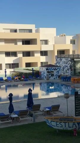 The video shows a swimming pool at a resort.  The pool is surrounded by lounge chairs with blue umbrellas.  In the background is a light-colored multi-story building with balconies.  The pool water is calm and reflects the blue sky and surrounding buildings.  Some people are lounging on the chairs. There is a small decorative boat on the lawn in the foreground. The weather appears sunny and clear.