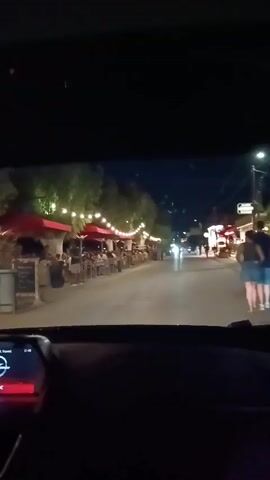 The video, taken from the perspective of a moving car at night, shows a street lined with outdoor dining establishments. String lights are visible above the restaurants, casting a warm glow. People are seated at tables, eating and socializing. Some of the tables are covered with red umbrellas. There are trees along the sidewalk, and the street is illuminated by streetlights and the lights from the restaurants. A person appears to be walking in the distance, and another person is visible pushing a stroller. The car appears to be driving down the street, passing by these establishments. The interior of the car is dark, with some dashboard lights visible. The overall atmosphere is lively and suggests a busy evening at an outdoor dining area.