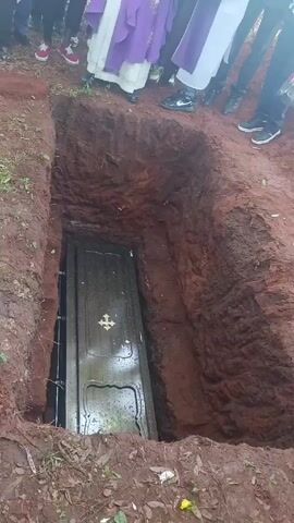 The video shows a burial. A dark coffin with a gold cross on top rests at the bottom of a rectangular hole dug into the red earth. Around the edge of the grave stand several people, including at least two clergy members wearing purple and white robes. Some people hold flowers. The ground is wet and muddy.