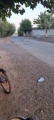 The video begins with a close-up of a bicycle wheel and the gravelly side of a road. As the camera pans to the right, a person is seen standing near a small outdoor kiosk. Further down the road, a child on a bicycle with a purple helmet rides away from the camera. The camera then pans to show a view of a street with houses and trees on either side. In the distance, two figures are walking down the road. The video continues to pan, showing a fenced-off area with trees behind it. The scene appears to be a rural or semi-rural environment, possibly in a village or small town, with the time of day appearing to be late afternoon or early evening, based on the lighting.