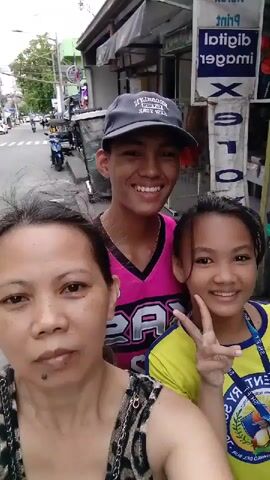 A woman is recording a selfie-style video with two children on a city street. The woman is in the foreground, wearing a sleeveless animal print top. Behind her to her right is a young girl with pigtails, wearing a yellow shirt and making a peace sign with her fingers. To the woman's left and partially behind her is a teenage boy wearing a pink basketball jersey and a baseball cap that says "Brooklyn New York". The video shakes slightly, suggesting the woman is holding the camera and perhaps moving. The background shows a typical urban street scene with buildings, power lines, and other vehicles, including a motorcycle with a sidecar. A sign for a print shop is visible behind them with reversed text. The overall impression is of a casual family outing.