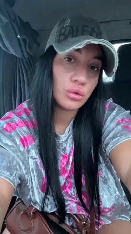 A woman with long, dark hair, wearing a grey baseball cap and a tie-dye shirt, is looking directly at the camera. Her lips are slightly parted, and she has a pouty expression. A brown bag is visible in the lower portion of the frame. The background appears to be the interior of a vehicle, with a curtain on the left side.
