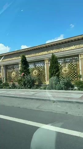 This video captures a drive through a street in Kasakh, Armenia, on a clear, sunny day. The camera, positioned inside a vehicle, offers a street-level view of various buildings and their surroundings.

The initial segment showcases an elaborate building with a white facade, adorned with gold accents and intricate moldings. A statue of a warrior in armor, holding a shield and spear, stands prominently on the rooftop. A large, ornate golden gate with a lion motif guards an entrance. Lush greenery, including a tall, slender evergreen tree and potted plants, adds to the sophisticated aesthetic.

As the drive progresses, the scenery shifts to more contemporary structures. One building features a modern grey exterior with awnings and signs displaying Armenian script. Several cars are parked in front of this establishment, suggesting it is a commercial space, possibly a restaurant or shop.

Further along, another building with a striking red and black geometric design on its facade comes into view. This building also appears to be a commercial establishment, with signage that includes the word "Cafe" and more Armenian text. Cars are again visible, parked along the street. The presence of road markings and sidewalks indicates a typical urban or suburban street environment. The overall impression is one of a lively daytime scene in a populated area.