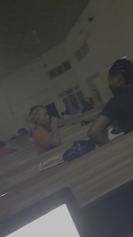 The video shows a low-light interior scene, possibly a lecture hall or classroom, where people are seated at desks. The camera view is unsteady, moving from a close up of two women talking, to a wider shot showing other people seated in rows, and then zooms in on a table surface with a phone on it. The overall lighting is dim, making it difficult to discern fine details. The room appears to have windows with a barred design. The women are talking, but the audio is inaudible, so the content of their conversation is unknown. The camera movements are jerky and not well-framed.
