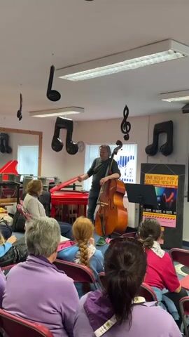 A man stands with a double bass, his left hand resting on the instrument. He is wearing a dark green long-sleeved shirt, blue jeans, and green sneakers. He is facing a group of people seated in chairs, their backs to the camera. The room is decorated with black musical note balloons and a treble clef ornament hanging from the ceiling. There is a red piano and a sheet music stand in the background. A poster with text and an image of a stage with spotlights is visible on the right side of the room. The lighting appears to be fluorescent.