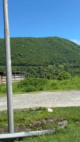 The video presents a drive-by view through a rural or semi-rural landscape in Achadara, Abkhazia, under a clear blue sky. Lush green, forested mountains form a prominent backdrop. Along the roadside, a series of structures are visible. These include several dilapidated or abandoned buildings made of concrete and brick, many appearing to have missing roofs, broken windows, or unfinished construction. One structure has a rusty tank on its roof, and a blue shipping container is situated near another. The road itself appears to be a mix of dirt and gravel, flanked by green grass and weeds. A metal guardrail runs parallel to the road. Further into the scene, a more modern-looking building with a dark roof can be glimpsed behind the older structures. A white sign, partially obscured by dense green vines, features some visible Cyrillic text, potentially including "ВИНОГОР" or similar, and "ЧАЧА," suggesting a possible connection to local wines or spirits. A triangular road sign with an arrow is also briefly seen. The scene depicts a sunny day with natural landscapes interspersed with human structures, some of which show signs of disuse.