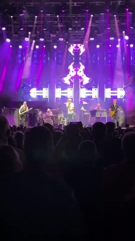 This is a video of a live concert performance. A band is on stage, with a lead singer in the center, a guitarist to his left, and another guitarist to his right. A drummer is visible behind them, along with a keyboardist. The stage is brightly lit with purple and red lights, and a large screen behind the band displays abstract visuals. The audience is visible in the foreground, with many people holding up their phones to record the performance. The overall atmosphere appears to be energetic and engaging.
