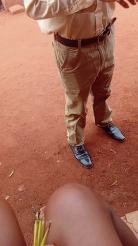 A man in a light beige button-down shirt, brown pants, and black shoes stands outside on a reddish-brown dirt surface. He is holding a small stack of money in his hand and speaking to the camera. The background shows a house with pinkish-red walls and a yellow door. There is a jerrycan, a basket, and what appears to be a set of stairs visible. The man is gesturing with his hands as he speaks. A leg is visible in the foreground.