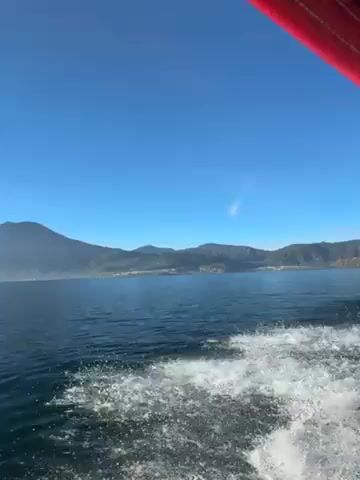 The video shows a view from a moving boat on a lake, with a mountain visible in the distance under a bright blue sky. The camera appears to be positioned at the front of the boat, capturing the water surface and the white wake being created by the boat's movement. The water has a dark blue hue and the mountain in the background is hazy. There's no people visible in the frame. The sun's reflection can be seen over the horizon.
