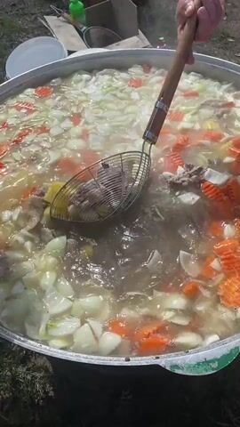 A high-angle, outdoor shot shows a large, metal pot filled with a boiling liquid containing potatoes, onions, carrots, and pieces of meat. An individual, wearing a long-sleeved garment, is adding potatoes by hand into the pot. Another individual is using a large slotted spoon with a wooden handle to stir the contents of the pot. The pot is placed on the ground in a grassy area. Other items, including plates and containers, are visible in the background, suggesting an outdoor cooking setup. Steam rises from the pot, indicating the high temperature of the cooking process.