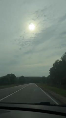 A dashcam view of a road through a landscape under a cloudy sky. The sun is visible through the clouds, appearing as a bright circle. Trees and foliage line the sides of the road, and in the distance, a faint tree line suggests a horizon. The road itself is paved and has white lane markings. A car is visible in the distance ahead, driving in the same direction. The video appears to be taken from inside a vehicle, with the dashboard visible at the bottom of the frame. The overall scene suggests a journey on a highway or rural road.