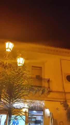 This is a vertical, medium-shot video of a building at night, lit by streetlights and fairy lights. The building is yellow and has several windows and balconies with wrought iron railings. In the foreground on the left, a palm frond is visible. A street lamp with three lights is also on the left side of the frame. The sky is dark and there is a small, faint light visible high in the sky. The video pans slightly to the right across the building. There are no people visible in the video.