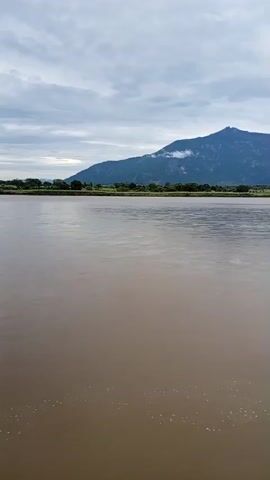 A wide shot shows a brown river with a mountain range in the background. The sky is cloudy. The camera is stationary.