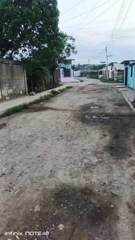 The video shows a view down a dirt road in a small town. The road is uneven with dark patches and puddles. On either side of the road are simple, mostly single-story houses. Many are painted in bright colors, such as blue and pink. Some houses have small yards or simple fences. There are also some concrete walls. A large tree stands to the left of the road, its branches reaching into the frame. Utility poles and wires run along the street. The sky is overcast. The perspective suggests the video was taken while walking down the road. The watermark "Infinix NOTE 40" is visible at the bottom of the screen.