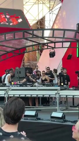 A group of people are on a stage in front of a large red and white geometric backdrop. Several people are standing behind DJ equipment, including a woman in the center wearing headphones and a black top. She is operating a DJ mixer. To her left, a man in a white tank top is holding a yellow cup and looking towards the DJ. To her right, a woman in a black tank top is watching. The stage has a metal frame structure above it with lighting equipment attached. In front of the stage, the backs of several people's heads are visible, suggesting an audience. The word "LOVERFEST" is printed repeatedly on a banner across the front of the DJ booth. The scene appears to be an outdoor music festival or event during daylight.