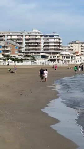 The video shows a beach scene.  A man and a woman walk together along the shoreline. The man is wearing a dark shirt and green shorts and carrying sandals.  The woman wears a white t-shirt and light shorts.  In the background, a large multi-story hotel building dominates the scene. Other people are scattered across the beach. The tide gently laps at the shore.