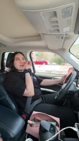 A woman with long dark hair, wearing a black t-shirt and black pants, is sitting in the driver's seat of a car. She is applying makeup to her face with a makeup brush. Her left hand is on the steering wheel, and her right hand is holding the makeup brush. A pink makeup bag is on the center console between the seats, containing various makeup products and a black water bottle. A can of soda is also visible on the center console. The car appears to be moving, with trees and buildings visible through the windows.