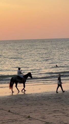 The video shows a man riding a horse on a beach at sunset. The man is wearing a white shirt and a dark hat, and he is holding a phone in his hand. The horse is walking along the wet sand near the water's edge. In the background, the ocean is calm, and the sky is a mix of orange and pink. Two people are walking on the beach, one wearing a straw hat and another wearing a colorful shirt. There is also at least one swimmer visible in the ocean. The beach appears sandy and somewhat reflective from the water.