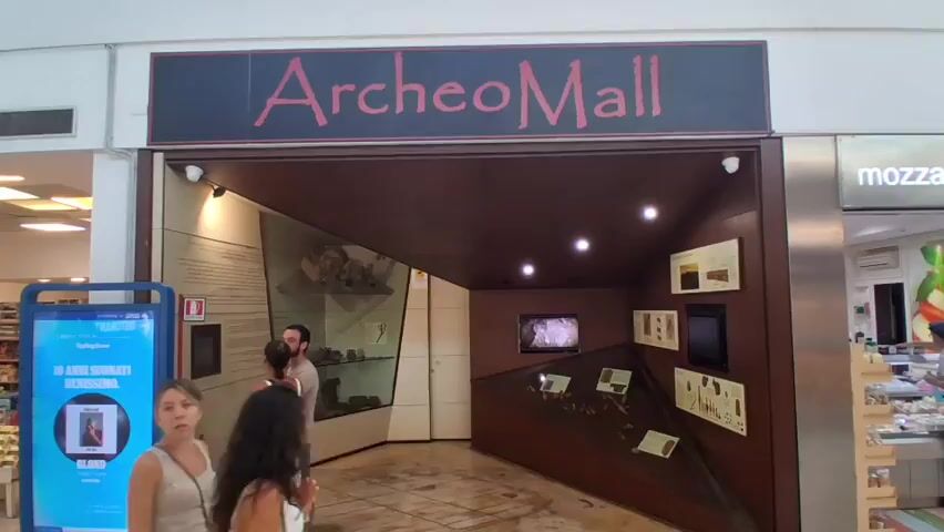 The video shows the exterior of a store called "ArcheoMall". There are people standing outside the store entrance, looking in. Inside the store, there are displays of artifacts and a video playing on a screen. A digital kiosk is also visible outside the store. The environment appears to be a shopping mall with tiled floors and lighting.