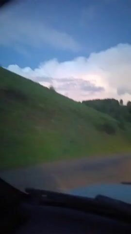 The video shows a first-person perspective from inside a vehicle, driving on a paved road. The sun is setting, casting a warm glow on the road ahead, and illuminating a white line down the center. Lush green hills with scattered trees line the sides of the road, and the sky above is a mix of blue and white clouds. The vehicle's headlights are on, casting a bright beam onto the road. The camera is focused on the road ahead, with the dashboard of the vehicle visible at the bottom of the frame. The overall impression is one of a scenic drive through a rural landscape at dusk.