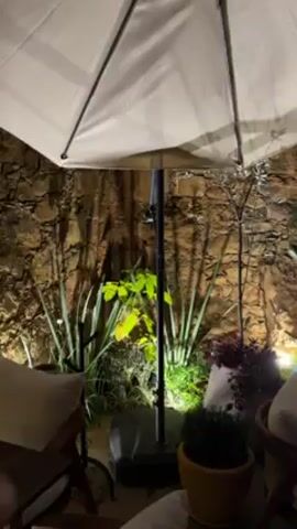 The video shows an outdoor seating area at night, likely a restaurant or bar.  There are several tables and chairs arranged under large umbrellas.  The area is lit with small, warm lights and there is a stone wall with plants behind the seating. No people are visible.  The camera slowly pans across the scene.