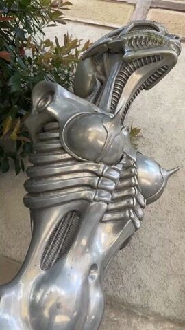 The video depicts a series of outdoor scenes in what appears to be a European town. It begins with a close-up of a metallic sculpture resembling a biomechanical figure. The subsequent shots show architectural details of buildings, including windows with shutters, arched doorways, and stone stairs, followed by a courtyard where a person is sitting on a couch, and a couple walking in front of a restaurant. The video concludes with a park-like area, capturing people relaxing in outdoor seating under a partly cloudy, blue sky.