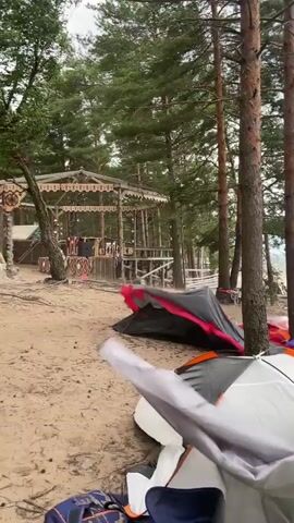 This video shows a campsite near a body of water, likely a lake or sea, with tall trees. Several tents are set up on the sandy ground. A person in a striped shirt and shorts is seen near some tents, appearing to be setting them up or organizing the campsite. Another person in a white shirt and pink shorts walks past a green tent. The weather appears overcast with a potential storm approaching, as indicated by dark clouds in the background. There are also some structures and beach chairs visible in the distance. The overall impression is of a recreational activity taking place outdoors.
